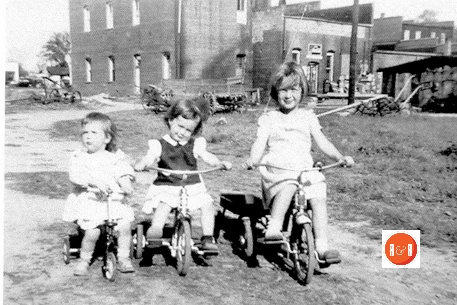 Betty, Susan and Judy Moore, daughters of George and Louise Moore, on their tricycles at the rear of the stores along North Main Street, Gray Court, S.C.